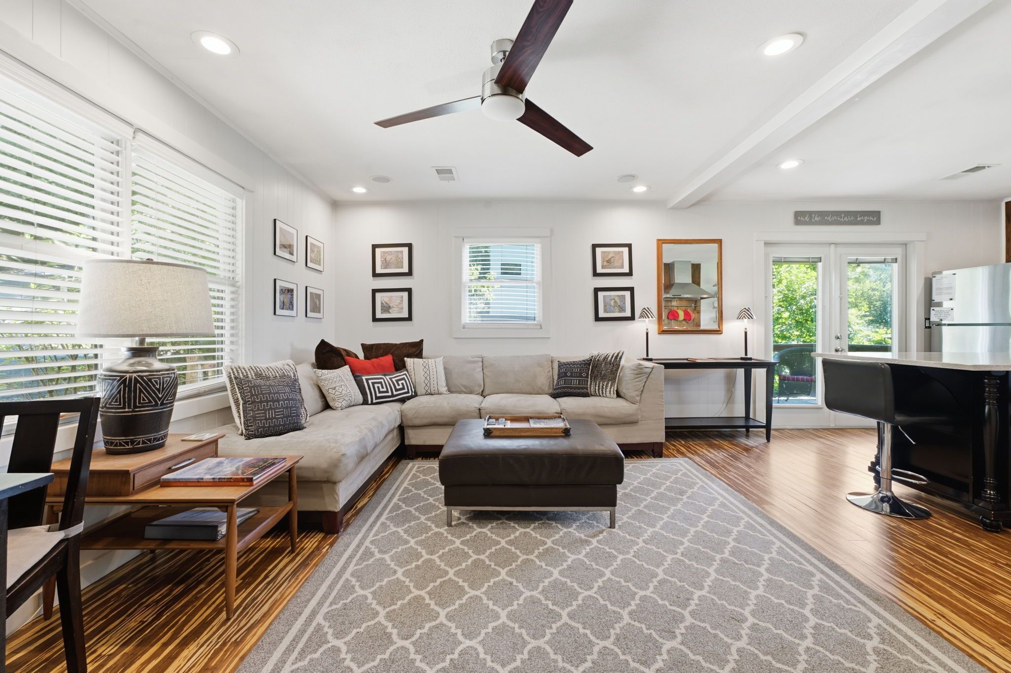 A modern living room with a sectional sofa, a coffee table, a ceiling fan, and a kitchen area with a refrigerator.