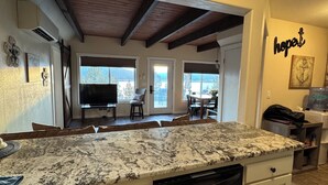 A kitchen with a granite countertop, white cabinets, and a dishwasher. There is a dining area with a table and chairs visible through an open door.