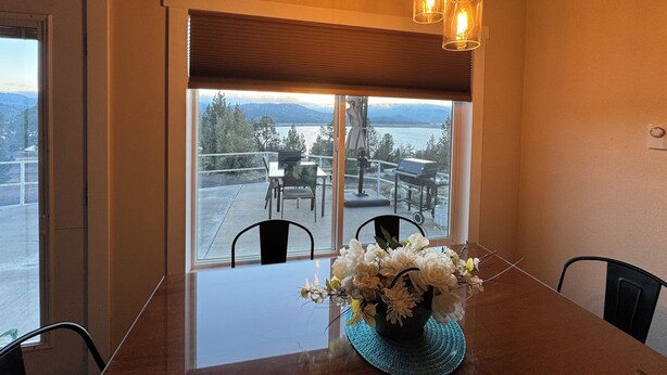 A dining area with a table, chairs, a vase of flowers, and a view of a lake and mountains through a large window.