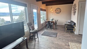 A spacious living area with a large TV, wooden flooring, a dining table with chairs, and a view of the outdoors through large windows.
