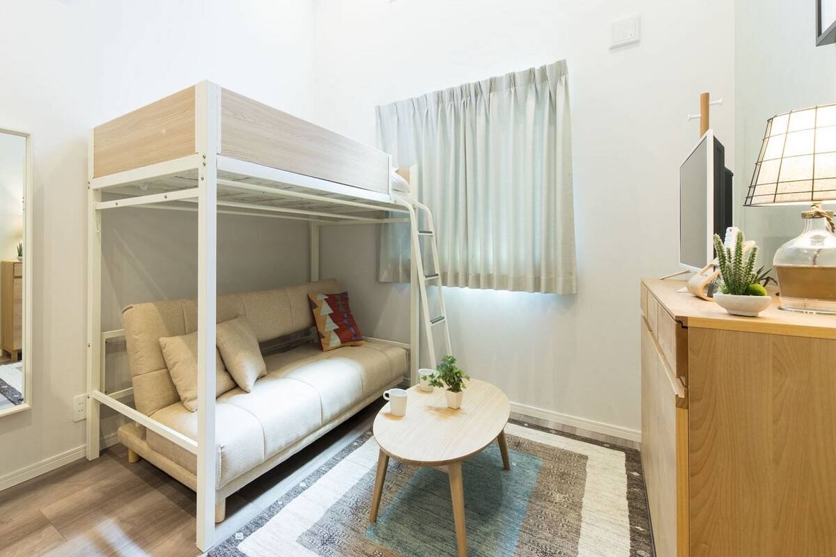A compact room with a bunk bed, a small table with a lamp, a mirror, a refrigerator, and a window with curtains.