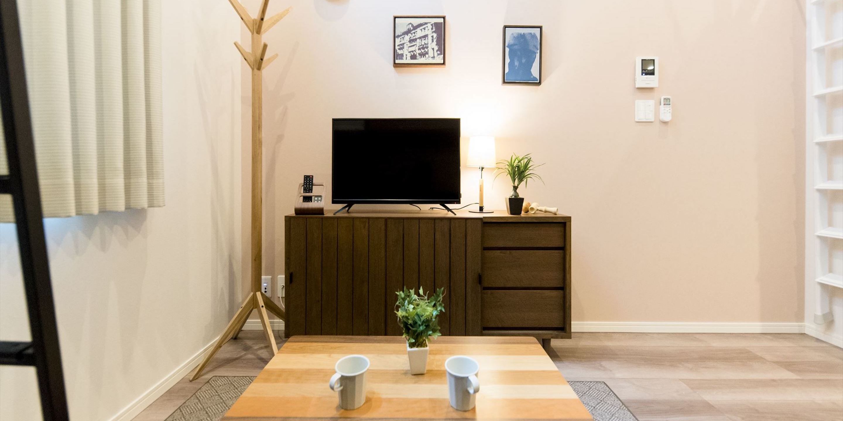 Een woonkamer met een televisie op een houten kast, een klein tafeltje met twee mokken, een plant en ingelijste foto's aan de muur.