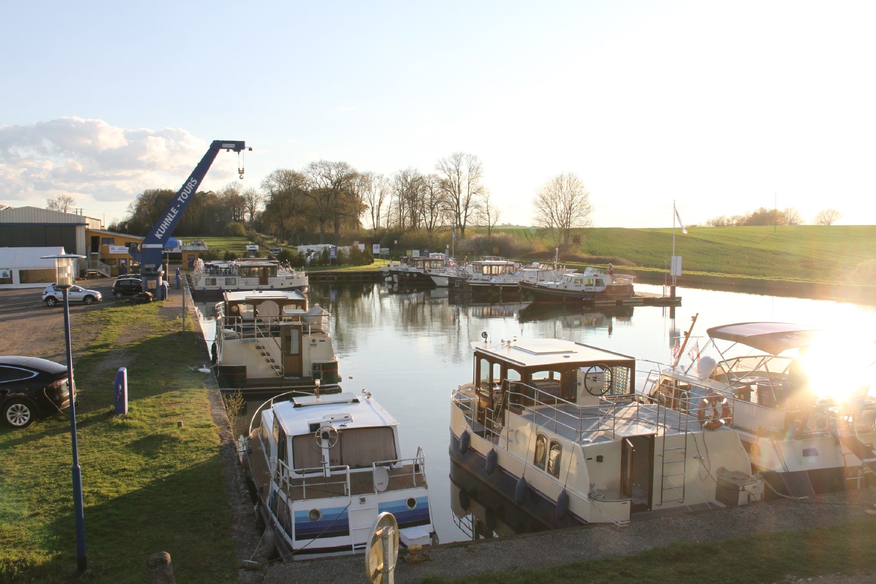 A marina with several boats docked, a crane, and a grassy area with a building in the background.