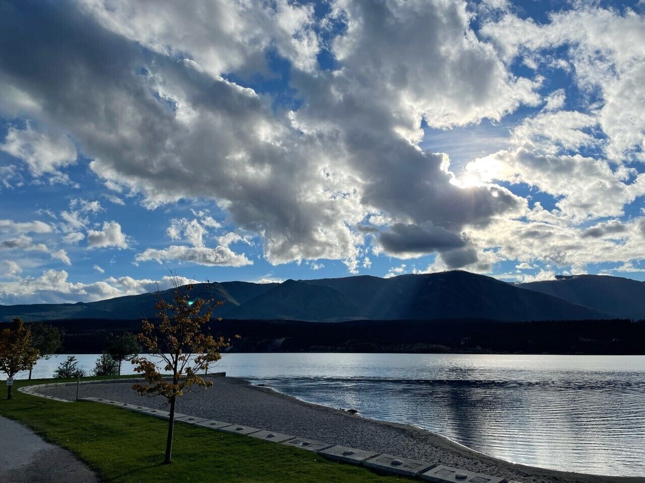 A lakeside view with mountains in the background, a paved path, and a few trees.