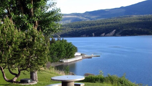 A picnic table with benches in a grassy area near a lake, surrounded by trees and a distant mountain range. - Private beach and lake access at Windermere Lake. NEW POOL AND HOT TUB NOW OPEN! (Windermere)