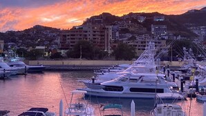 Un puerto deportivo con varios yates amarrados, un atardecer de fondo y una colina con edificios.