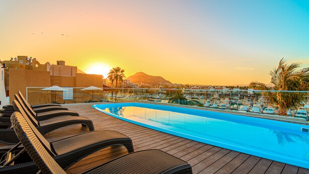 Una piscina en la azotea con sillones de descanso, vistas a un puerto deportivo y una montaña al fondo.