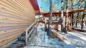 A wooden cabin with a staircase leading to a deck, surrounded by trees and snow.