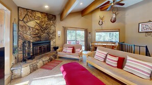 A cozy living room with a stone fireplace, a sofa with striped cushions, a red bench, and a dining area with a view of the outdoors.