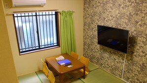 A room with tatami flooring, a wooden table with chairs, a television on the wall, and a window with metal bars and green curtains. - Shirakabanoyado-keigetsu (Osaka)