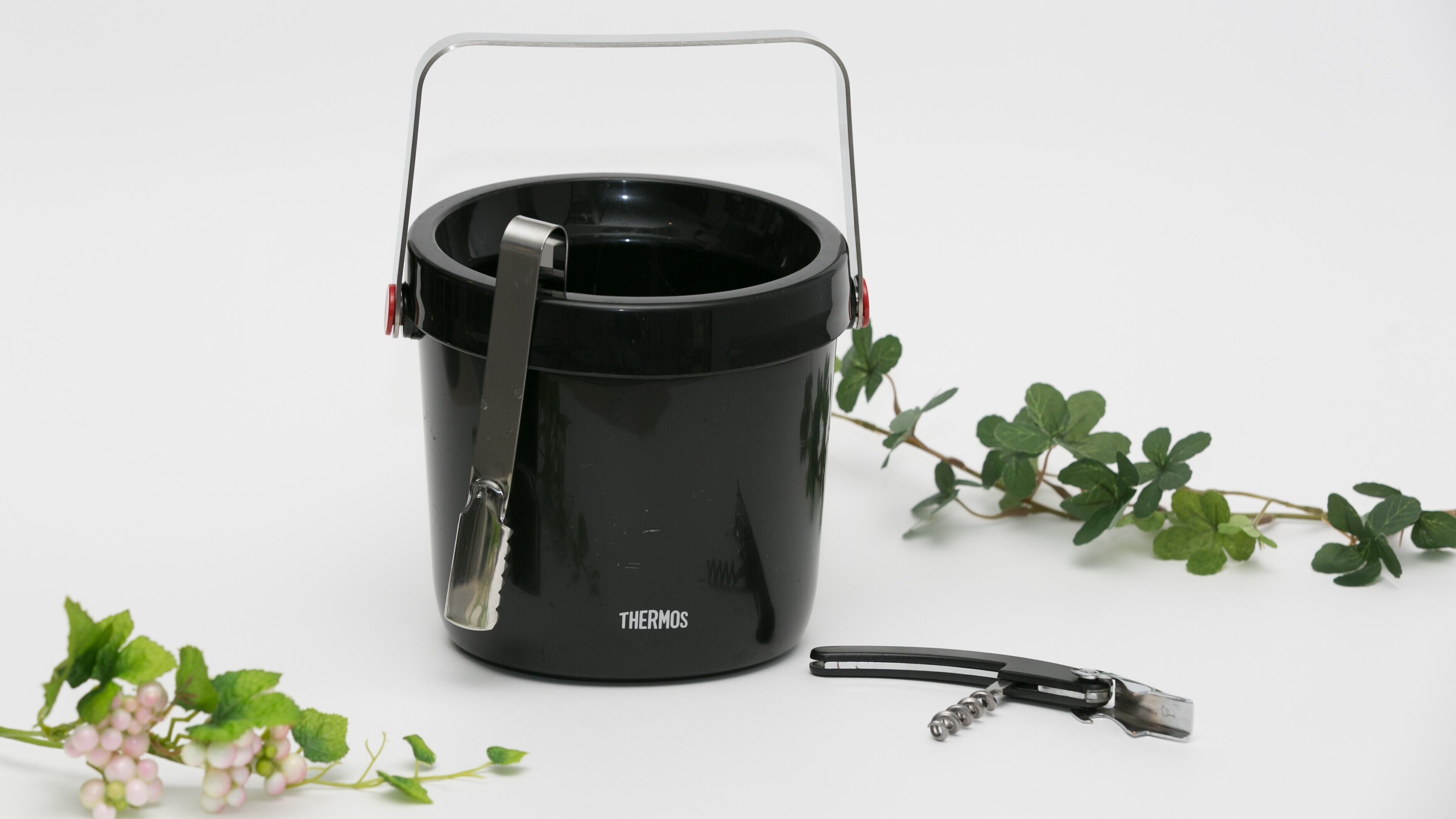 A black thermos with a handle and a strap, placed on a white surface with a plant and a metal object nearby.