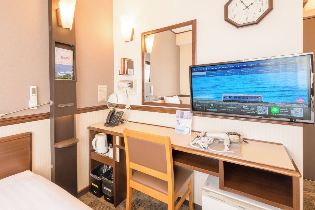 A hotel room with a bed, a desk with a computer, a telephone, a mirror, and a clock on the wall.