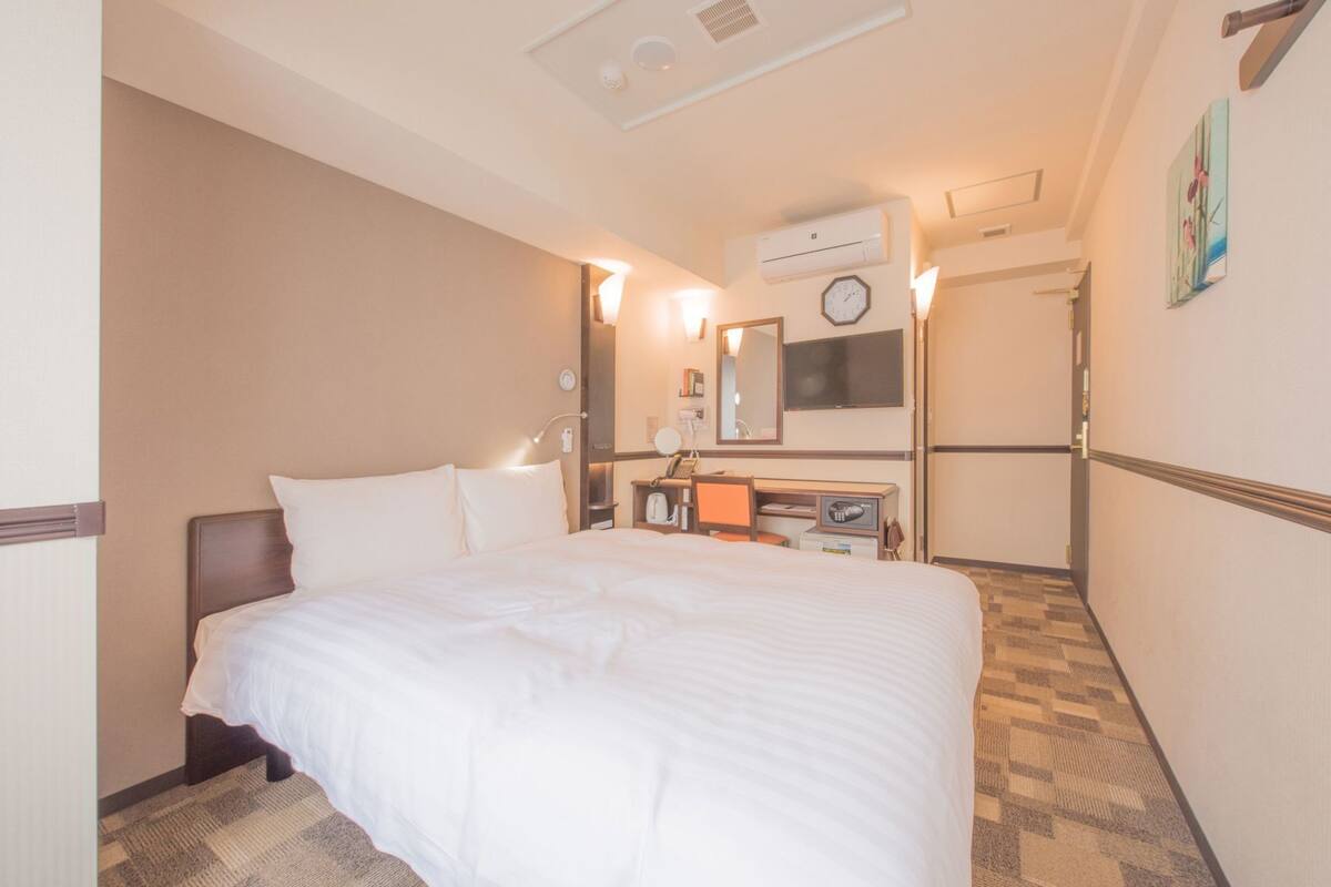 A hotel room with a large bed, a desk with a chair, a mirror, a clock, and an air conditioning unit.