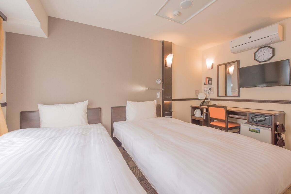 A hotel room with two beds, a desk with a chair, a mirror, a clock, and an air conditioning unit.