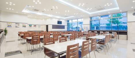 A spacious cafeteria with multiple tables and chairs, large windows, and a modern ceiling design.
