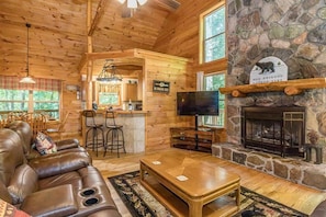 A log cabin interior with a leather sofa, a stone fireplace, a TV, and a kitchen area. - DELUXE FAMILY CABIN | HOT TUB | GAME ROOM | (Cleveland)