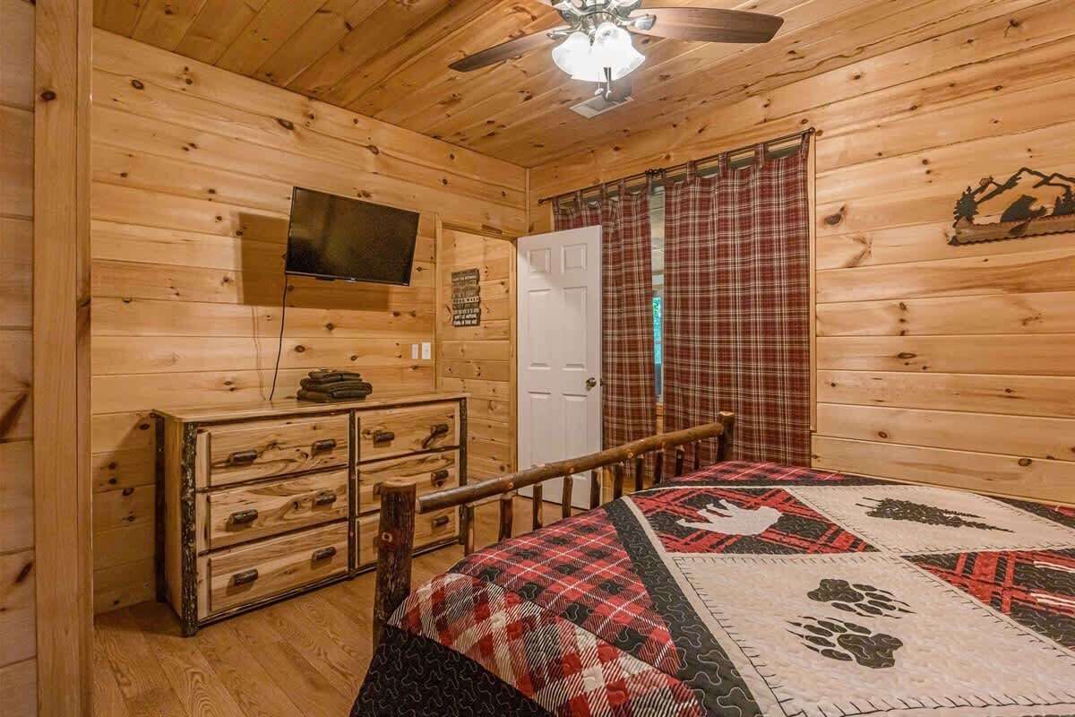 Ein Zimmer in einer Holzhütte mit einem Bett, einer Kommode, einem Fernseher und einem karierten Vorhang.