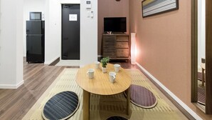 A compact living space with a dining area featuring a round table, four chairs, and a small potted plant. A black refrigerator and a wooden cabinet with a television are visible in the background. - Trip Pod Sumiyoshi A (Fukuoka)