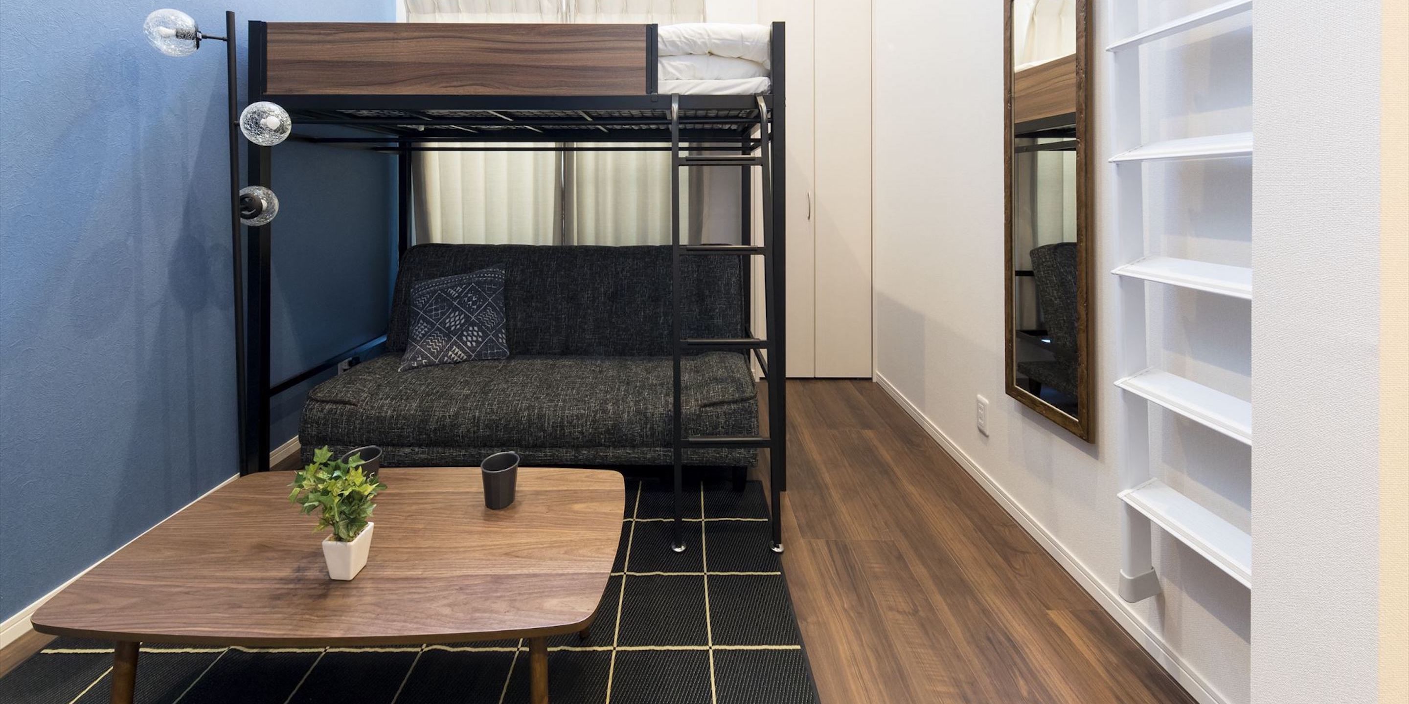 A compact room with a bunk bed, a small sofa, a table with a plant, and a window with blinds.