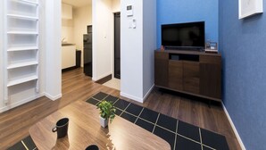 A modern living room with a wooden floor, a dining table with chairs, a TV on a wooden stand, and a blue accent wall. - Trip Pod Sumiyoshi A (Fukuoka)
