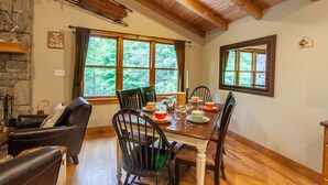 A cozy cabin interior with a dining table set for four, surrounded by wooden chairs, a fireplace, and large windows offering a view of the forest. - Cabin By Blue Ridge Parkway (Blowing Rock)