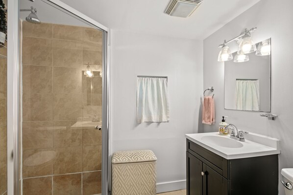 Une salle de bain avec une porte de douche en verre, un lavabo blanc avec un meuble sombre et un miroir avec éclairage intégré.