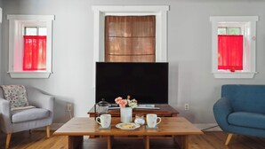 A living room with a wooden coffee table, a sofa, a TV, and a window with red curtains. - Cottage w/ Private Fenced Yard | Dogs OK (Wilmington)