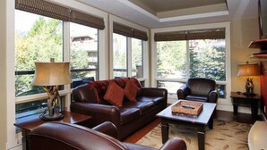 A living room with leather sofas, a coffee table, and a lamp. Large windows provide a view of the outdoors. - Mökki Mountain Suite - mountain views & private corner unit (Canmore)