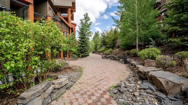 A paved pathway flanked by stone walls and greenery, leading to a building with wooden balconies and a clear sky. - Mökki Mountain Suite - mountain views & private corner unit (Canmore)