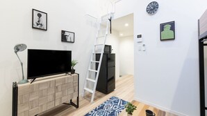 A modern living room with a flat-screen TV on a wooden TV stand, a white ladder, a black refrigerator, and a blue patterned rug. - Trip Pod Sumiyoshi C (Fukuoka)
