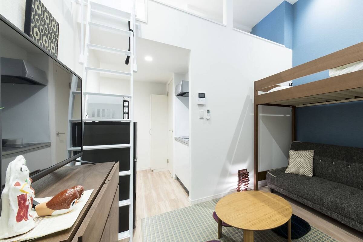 A bunk bed with a wooden frame and a sofa underneath, a small round table, and a kitchen area with a microwave and a stove.