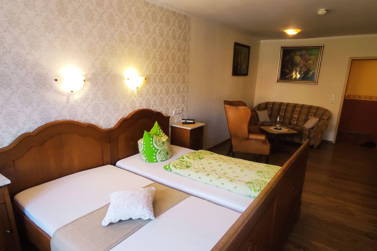 A hotel room with a wooden bed, a chair, a small table, and a painting on the wall.