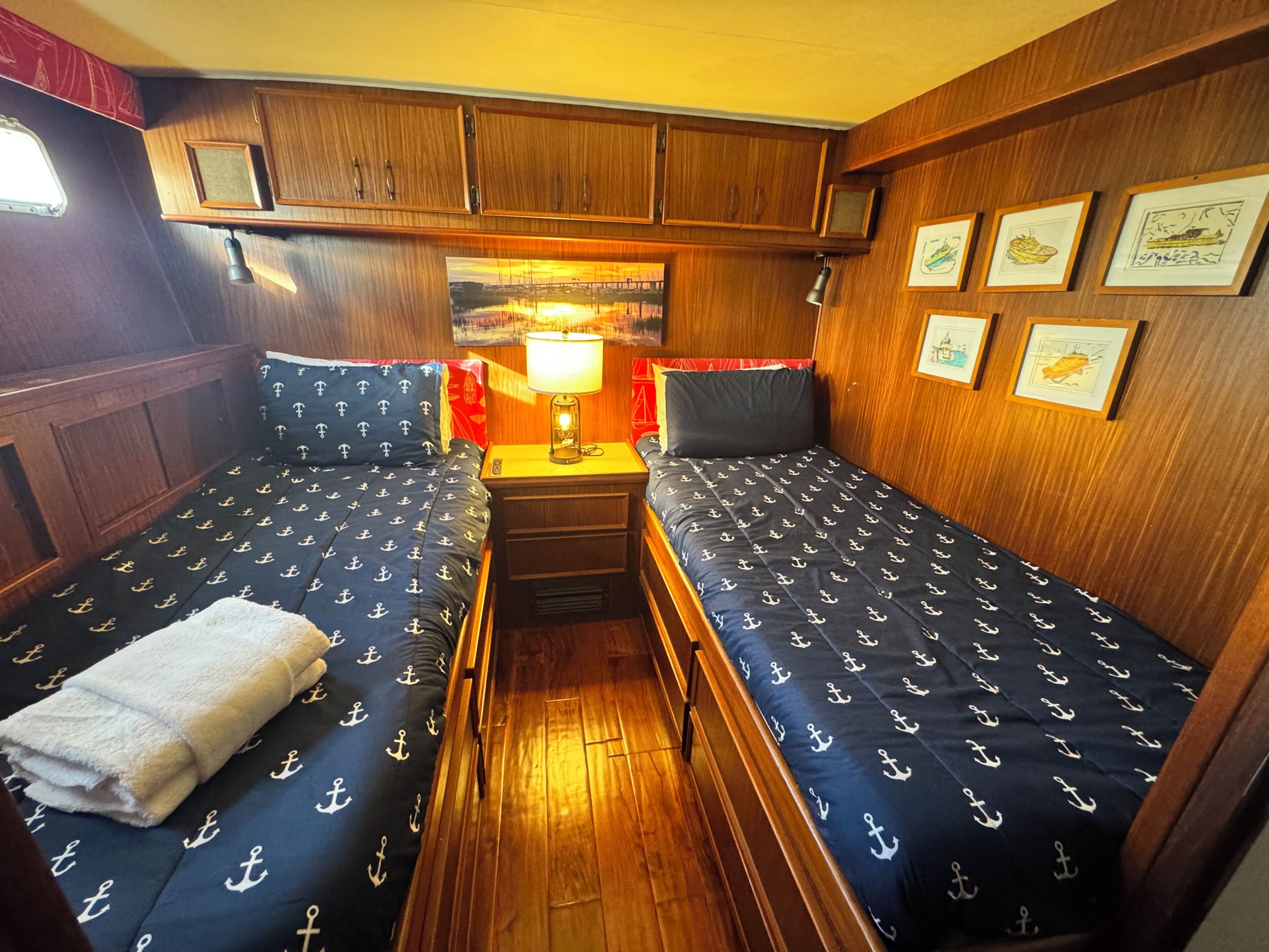 A cabin with two beds, a small table with a lamp, wooden walls, and framed pictures.