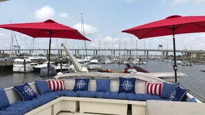 Un pont de bateau avec une rangée de sièges bleus rembourrés et des parasols rouges, donnant sur une marina où sont amarrés de nombreux bateaux, avec un pont en arrière-plan.