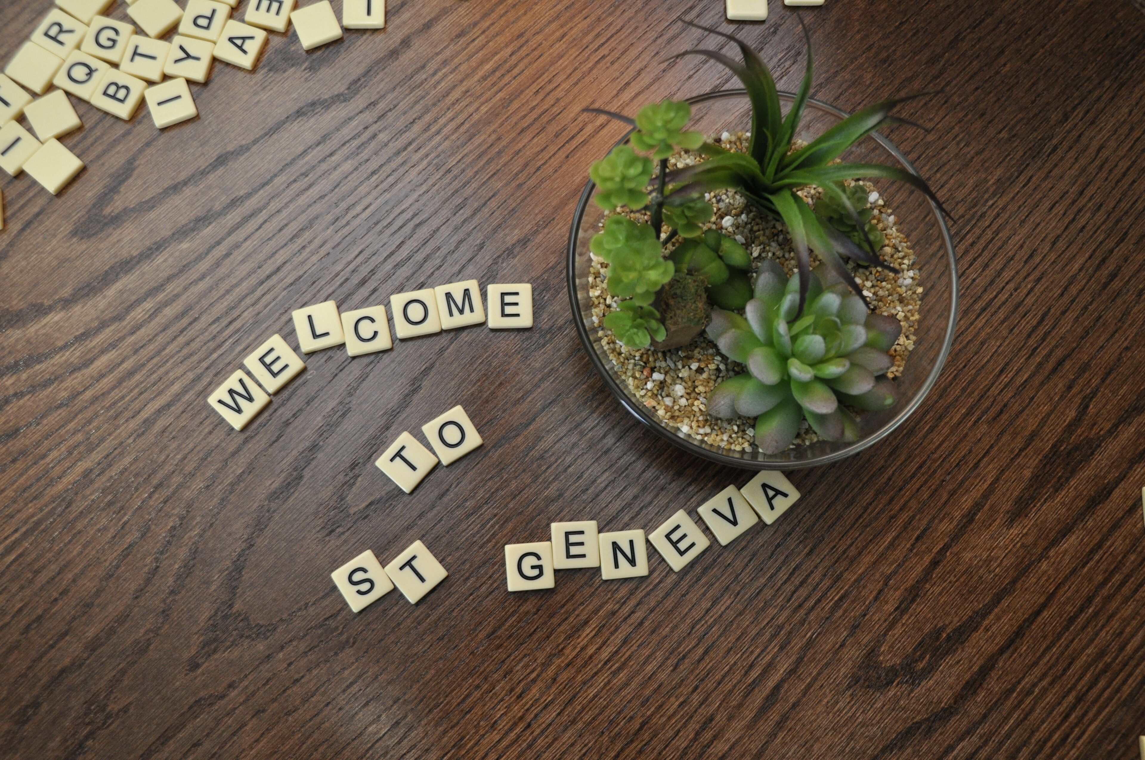 A welcome mat spelling "WELCOME TO ST GENEVA" on a wooden floor with scattered letter tiles.