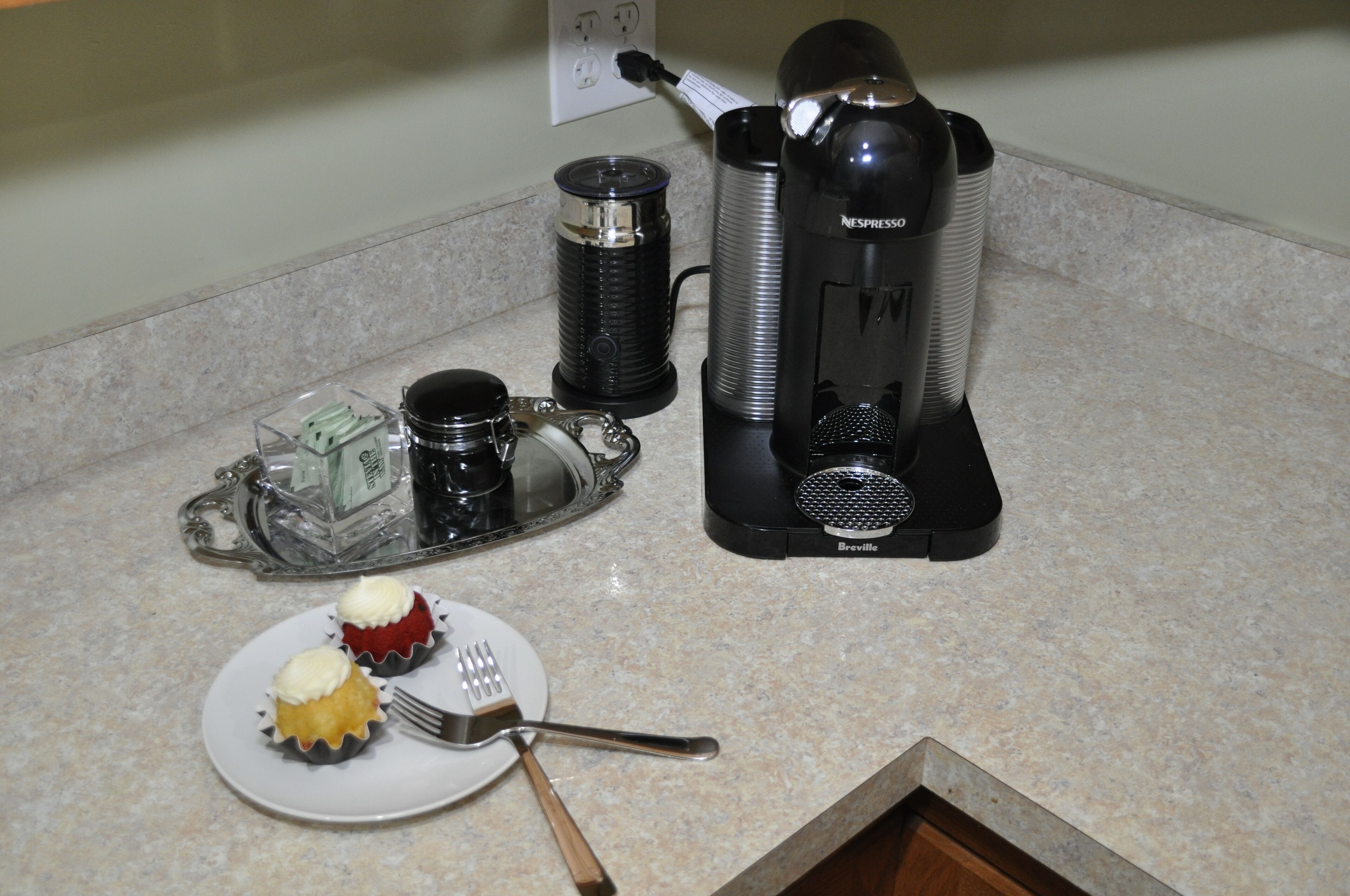 A kitchen counter with a coffee maker, a glass carafe, a metal pitcher, a plate with cupcakes, and a fork.