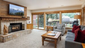 A living room with a fireplace, a television mounted above it, a sofa, a coffee table, and a view of the outdoors through large windows. - Montaneros 314 - 1 Bedroom Platinum (Vail)