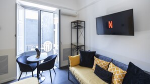 A modern living room with a sofa, two chairs, a TV mounted on the wall, and a balcony with a table and chairs.