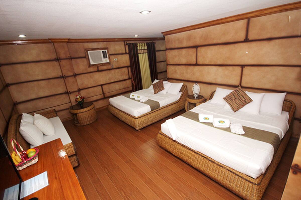 Une chambre d’hôtel avec un grand lit, un lit plus petit et un canapé. Le sol est en bois, il y a un téléviseur accroché au mur et une fenêtre avec des rideaux.