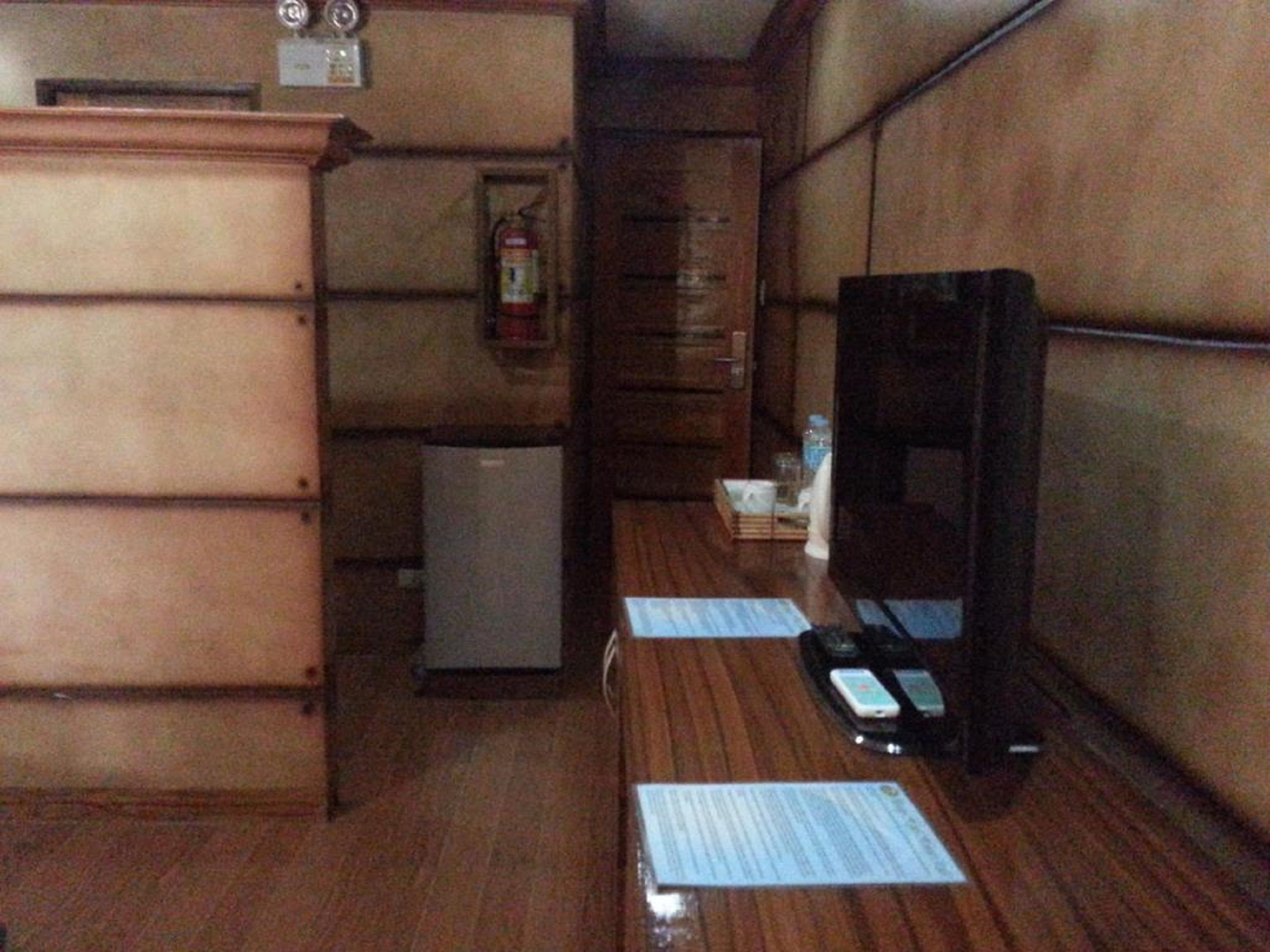Quarto compacto com escrivaninha de madeira, pequeno frigorífico, extintor de incêndio e televisão.