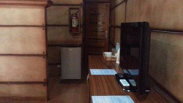 Quarto compacto com escrivaninha de madeira, pequeno frigorífico, extintor de incêndio e televisão.