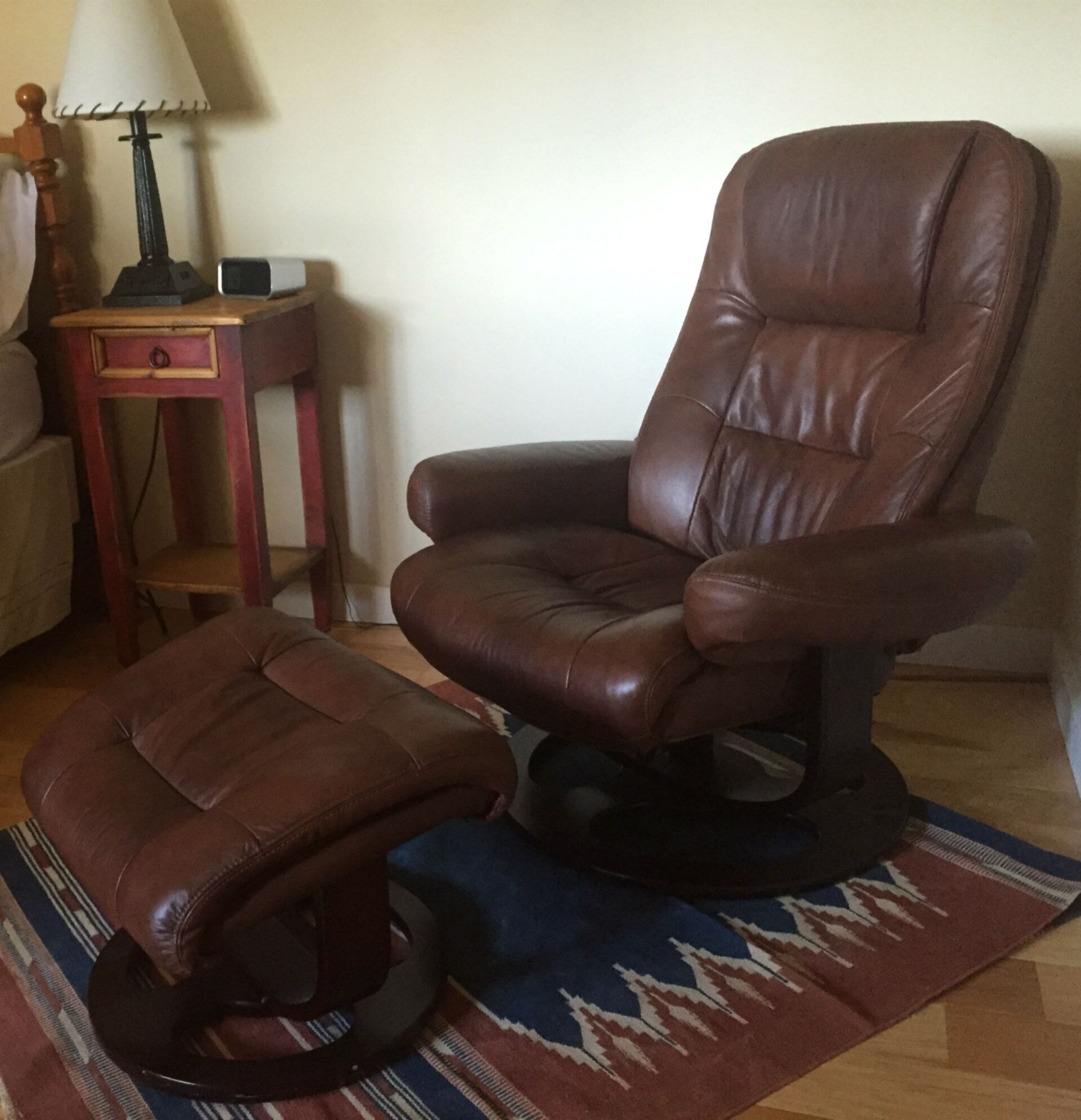 Un sillón reclinable de cuero marrón con su otomán a juego, una mesita de madera pequeña con una lámpara, y una alfombra estampada en el suelo.