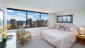 Une chambre d’hôtel dotée d’une grande fenêtre donnant sur la ville, d’un lit recouvert d’une couette beige, d’une table de chevet avec une lampe et d’un fauteuil en osier agrémenté d’une plante en pot.