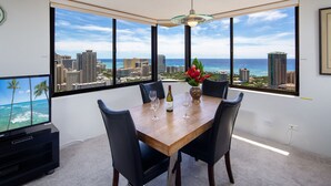 Un coin repas avec vue sur la ville, une table avec des chaises, un vase rempli de fleurs et un téléviseur diffusant des images d’un paysage tropical.