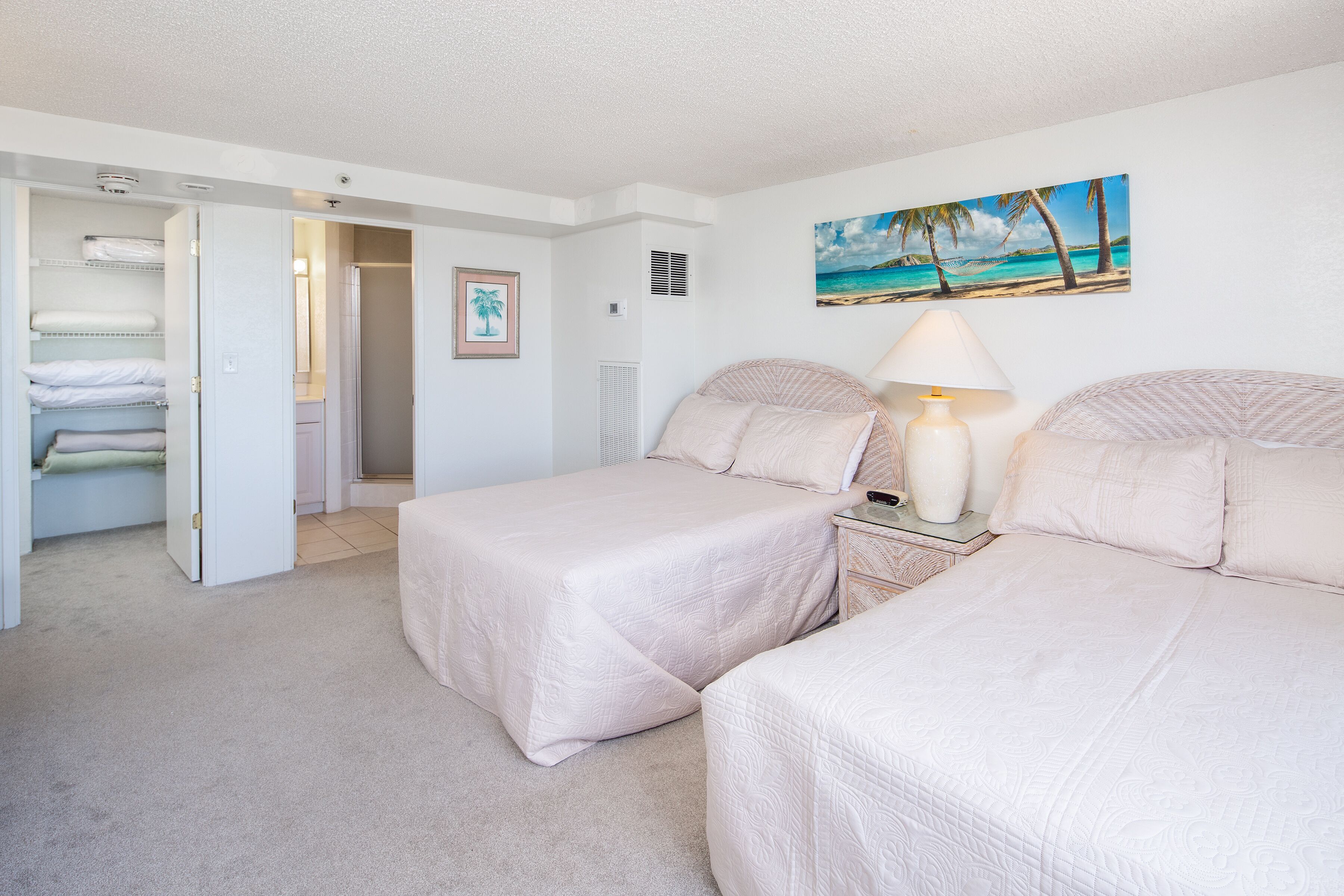 A bedroom with two beds, a nightstand with a lamp, a painting of a beach scene, and a closet with shelves.