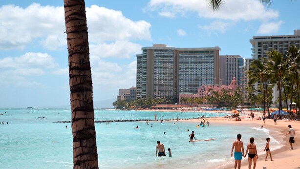 Une plage à l’eau turquoise et limpide, bordée de palmiers, avec de grands immeubles qui se dressent à l’arrière-plan.