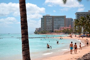 A beach with clear turquoise water, palm trees, and high-rise buildings in the background.
