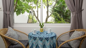 Een kamer met een raam, twee rieten stoelen, een klein rond tafeltje met een tafelkleed met patroon, en een potplant.