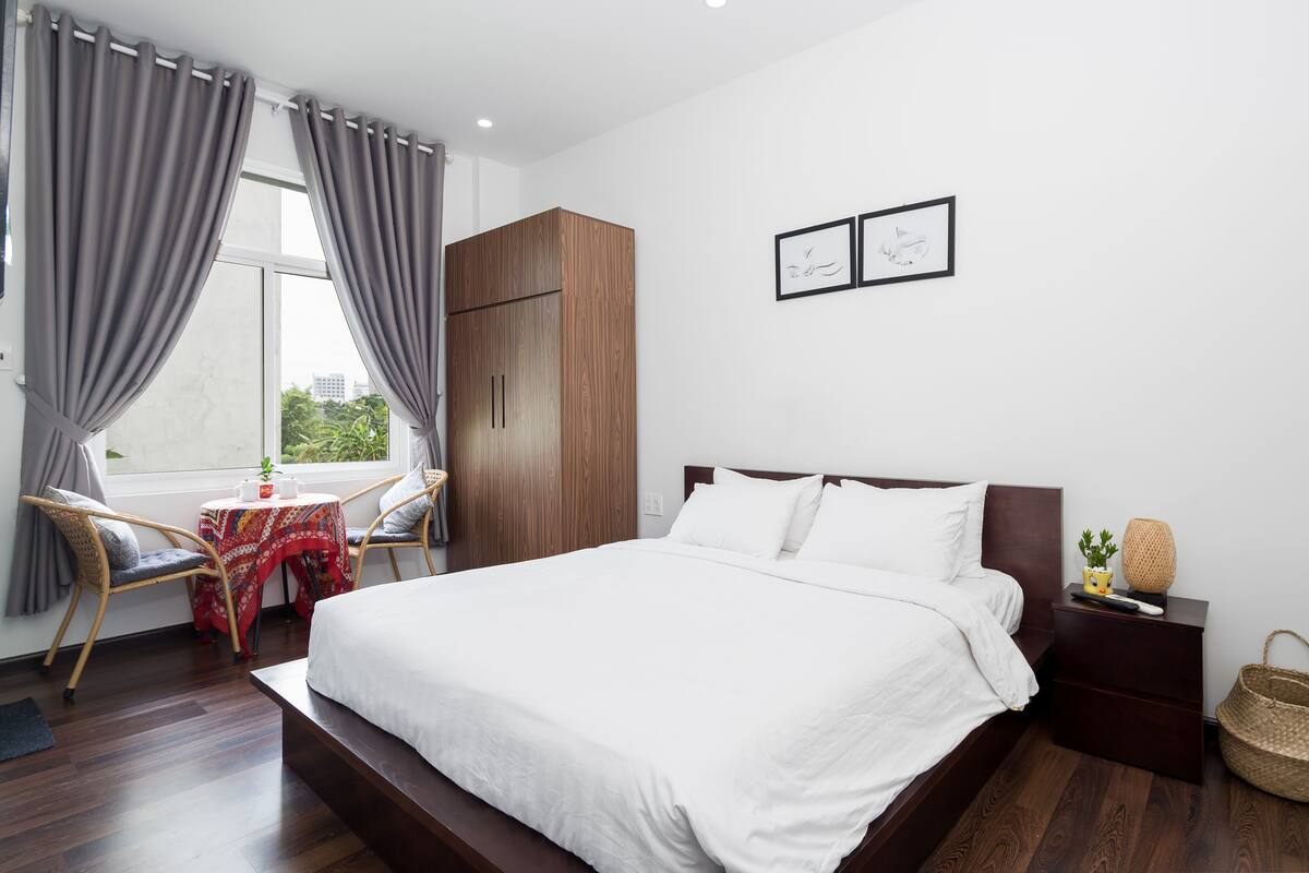 Ein Schlafzimmer mit einem großen Bett, einem Holzschrank, einem kleinen Tisch mit roter Tischdecke, Korbstühlen und einem Fenster mit Vorhängen.