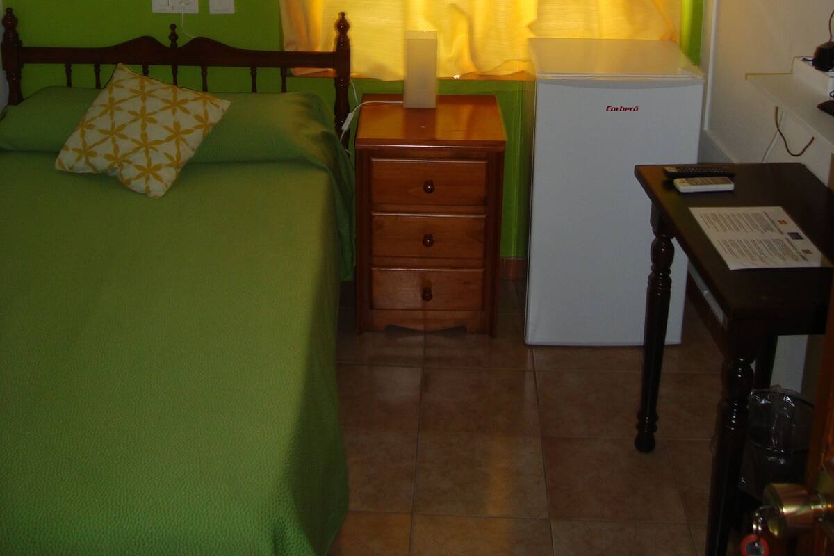 A bedroom with a bed, bedside table, lamp, and a small desk with a chair.